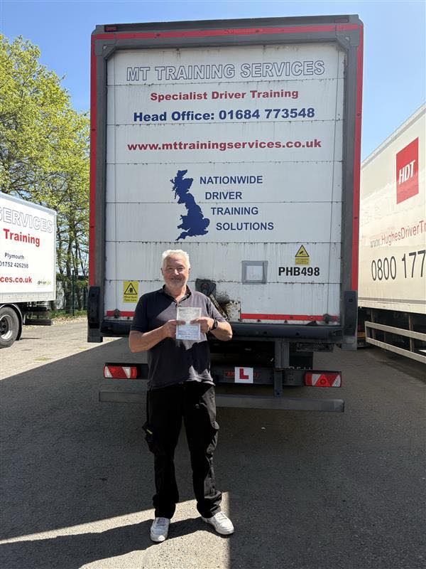 Male trainee driver holding pass certificate in front of HGV training vehicle after passing CE 3a test at MT Training Services Tortworth